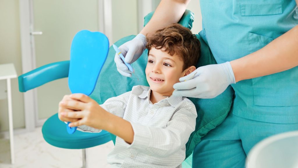 A child in a dentist chair, holding a blue mirror, with a dentist attending to him from behind.