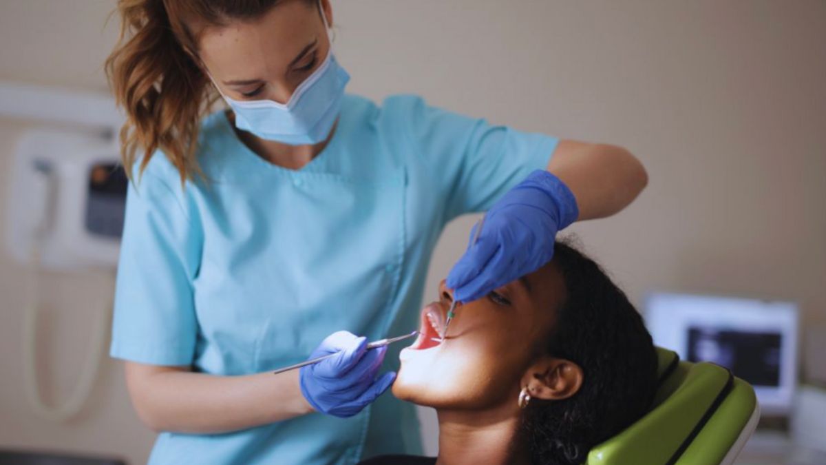 A dentist in a light blue uniform, face mask, and blue gloves examines a dark-haired woman's teeth as she reclines in a green chair.