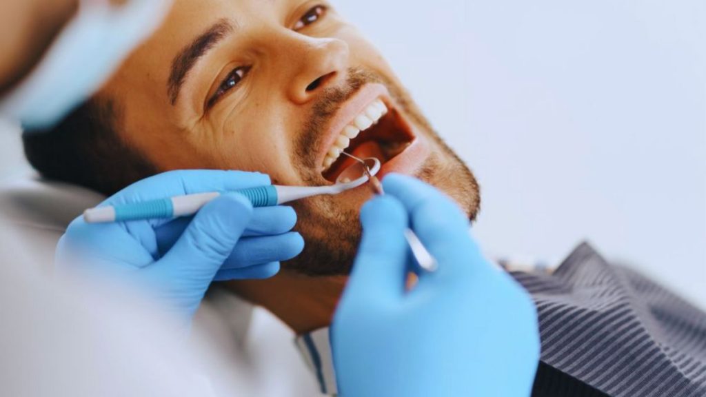 A dentist in blue gloves examines a patient's teeth in a dental chair.