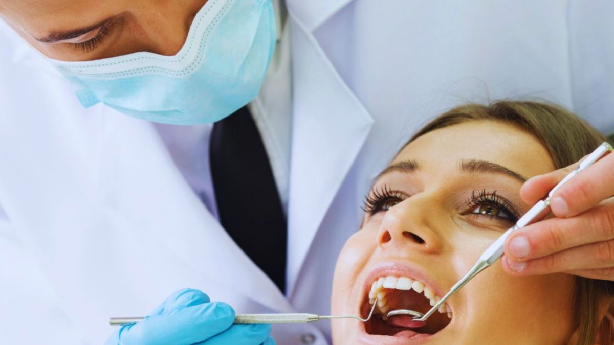 A dentist wearing a mask and gloves examines a smiling female patient's teeth using dental instruments in a clinical setting
