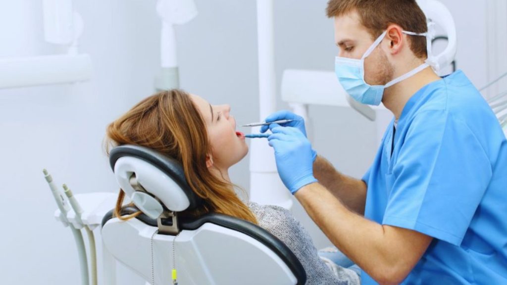 A dentist wearing blue scrubs, gloves, and a face mask is examining a female patient's teeth in a modern dental clinic.