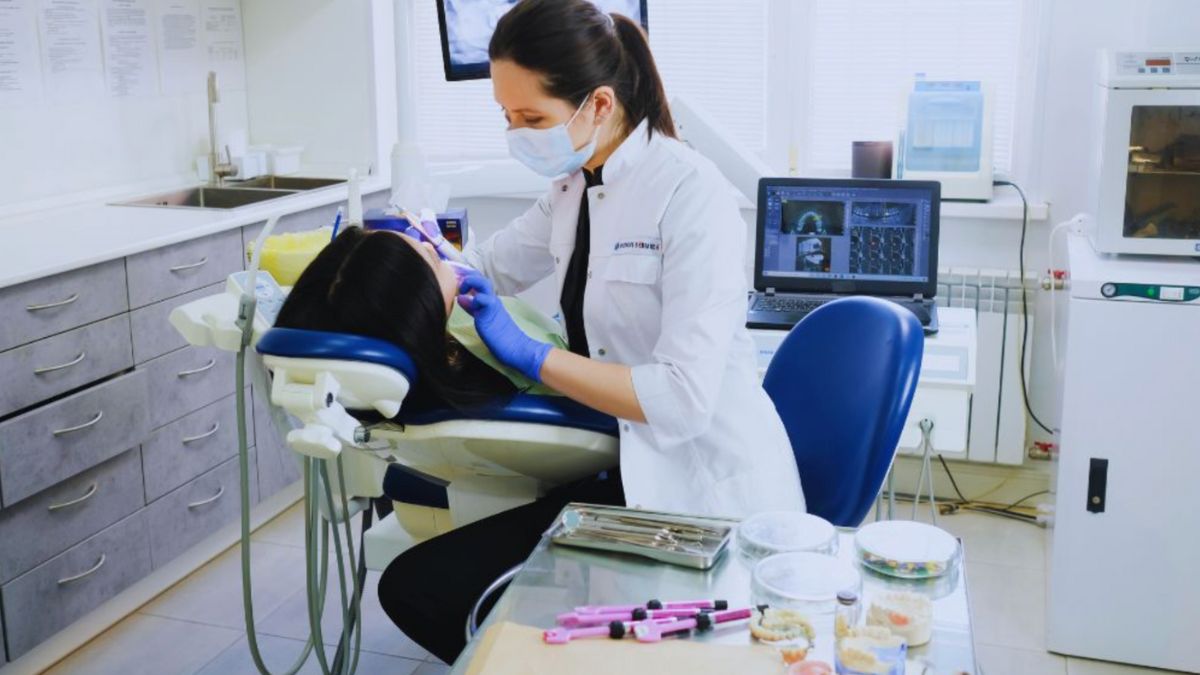 A dentist in a white coat, gloves, and a face mask treats a patient in a dental chair. The clinic features modern technology, including a computer displaying dental X-rays, sterilized tools, and well-organized medical supplies.