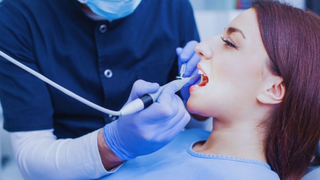 A dentist wearing a navy blue uniform, face mask, and blue gloves is performing a dental procedure on a female patient with long brown hair. The patient, wearing a light blue bib, has her mouth open while the dentist uses a dental scaler and suction tool.