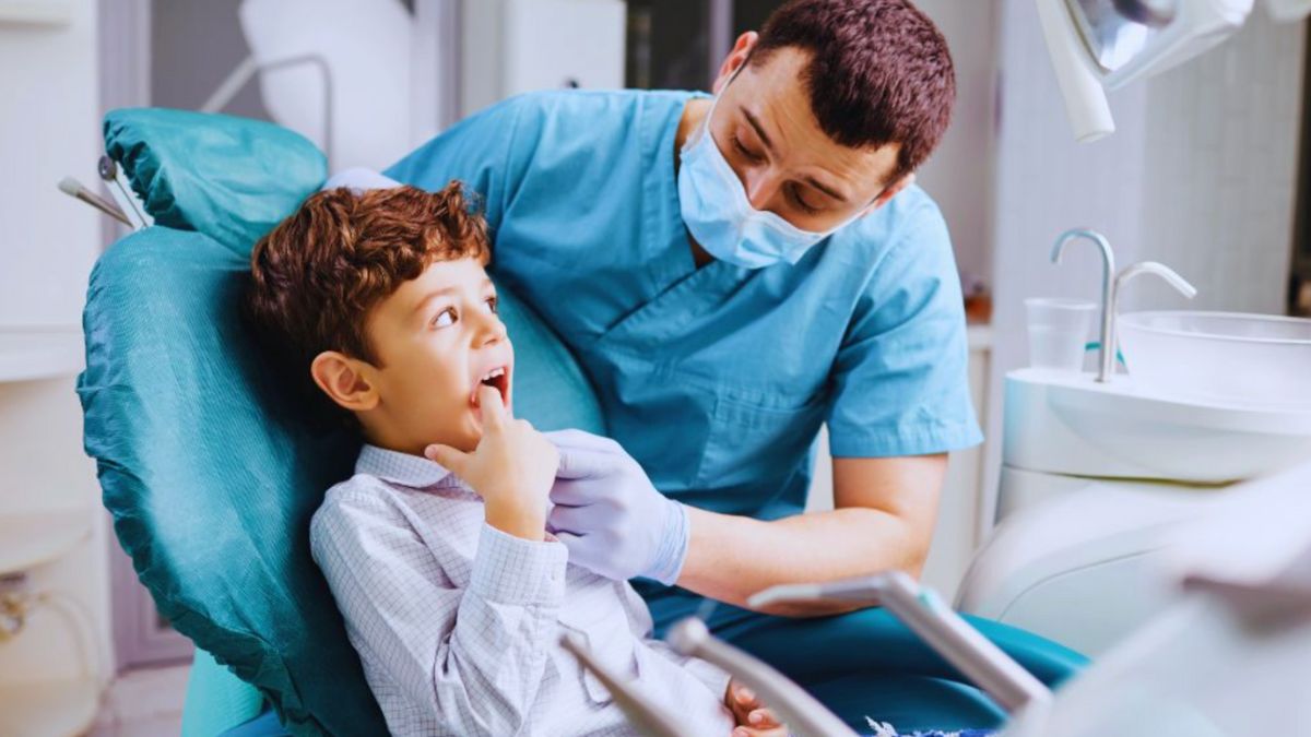 A young boy with curly brown hair sits in a dentist's chair, pointing at his teeth while looking at a male dentist. The dentist, wearing blue scrubs, a face mask, and gloves, attentively listens to the child with a friendly expression.