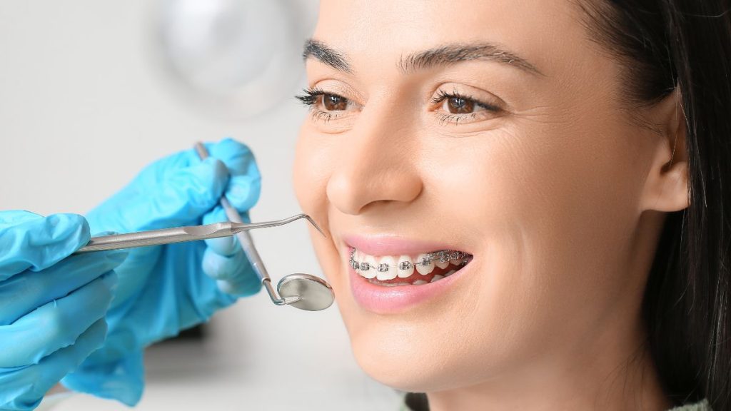 A smiling woman with braces sits in a dental chair as a dentist in blue gloves prepares to adjust her braces.
