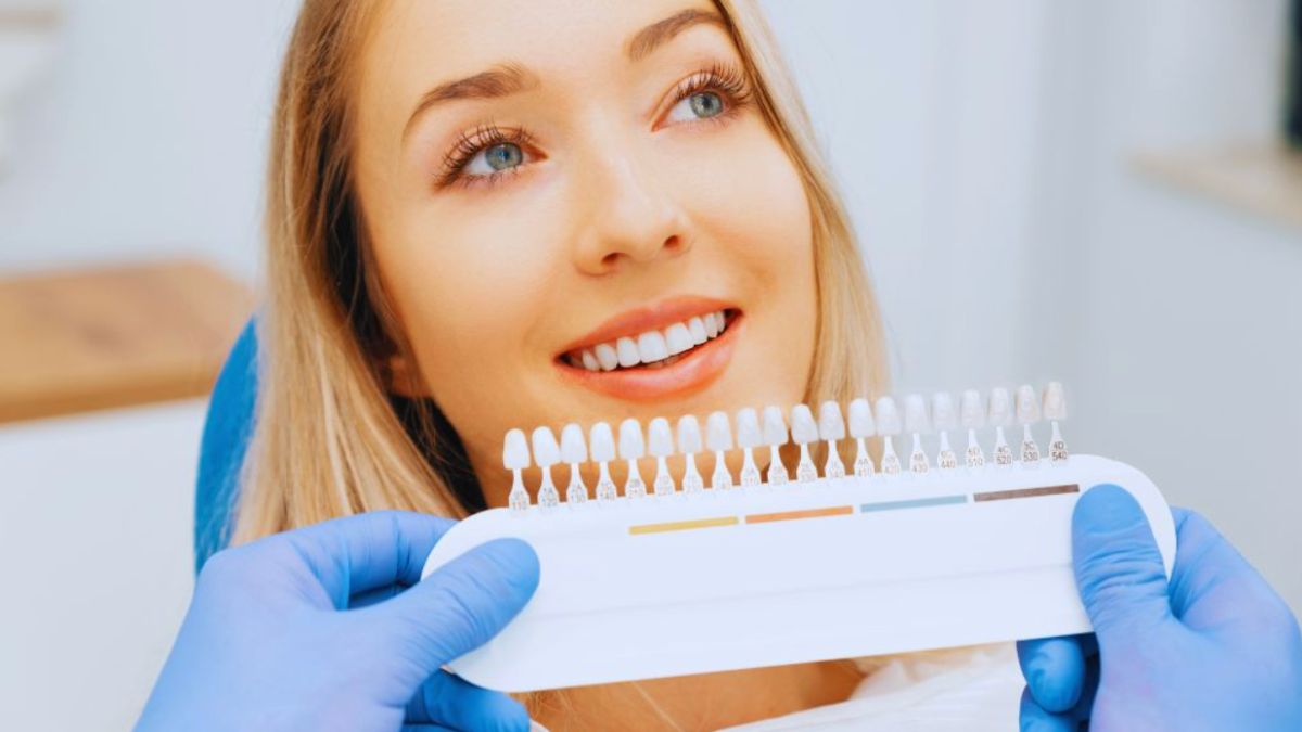 Woman smiling in a dental chair while a dentist holds up a tooth shade guide to match veneer color — Veneers Lake Worth, FL.