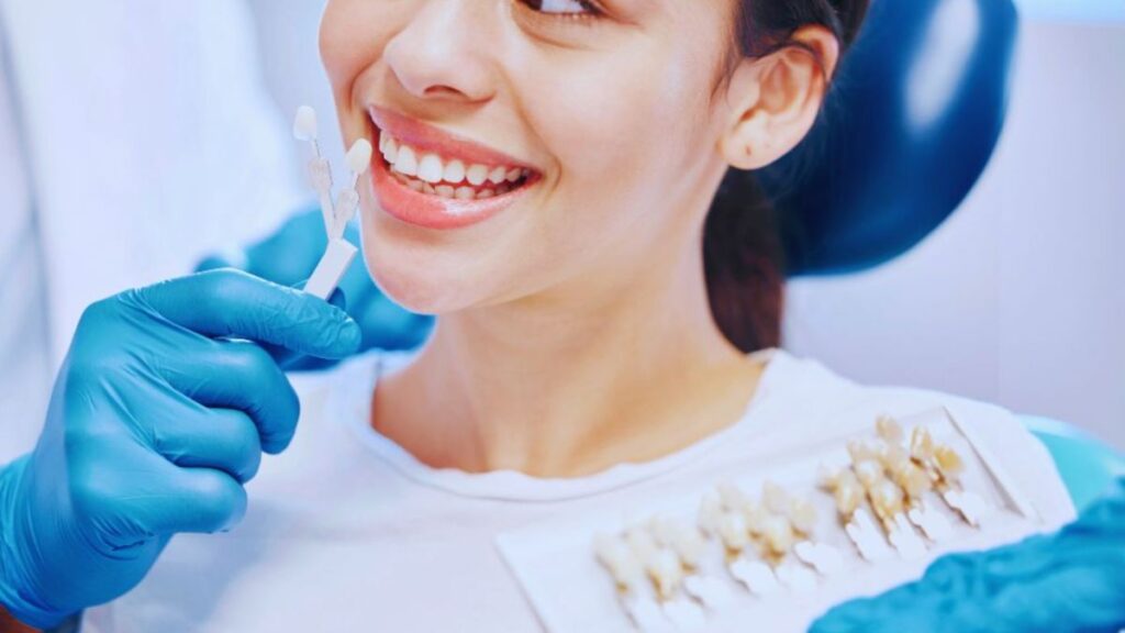 A dentist wearing blue gloves holds a shade guide next to a patient's teeth to select the best match for dental veneers, with various tooth-colored samples visible, taken in a clinical setting in Wellington, FL.